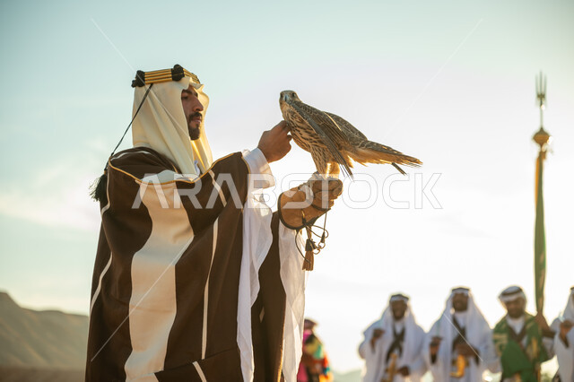 The day we started February 22, interest in raising and training birds, the status of falcons in Saudi history, the anniversary of the founding of the first Saudi state 1727 AD, a popular national occasion, a Saudi Gulf Arab man wearing a ghutra and a bisht carrying a falcon in his hand with expressions of pride, Al-Ardah Al-Najdi background