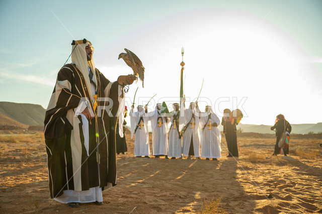 The status of falcons in Saudi history, the anniversary of the founding of the first Saudi state in 1727 AD, a popular national occasion, the day we started on February 22, interest in raising and training birds, a Saudi Gulf Arab man wearing a ghutra and a bisht carrying a falcon in his hand with expressions of pride, the background of the Najdi Ardah in the desert
