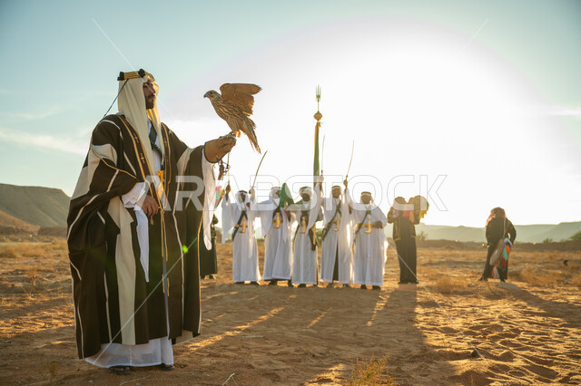 مكانة الصقور في التاريخ السعودي, ذكرى تاسيس الدوله السعوديه الاولى 1727 م, مناسبة شعبيه وطنيه, يوم بدينا 22 فبراير، الاهتمام بتربية الطيور و تدريبها, رجل عربي خليجي سعودي يرتدي الغترة والبشت يحمل على يده طائر الصقر بتعابير الفخر, خلفية العرضة النجدية في الصحراء