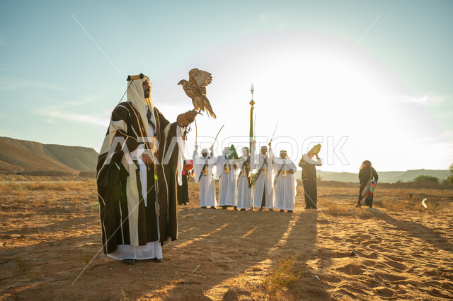 ذكرى تاسيس الدوله السعوديه الاولى 1727 م, مناسبة شعبيه وطنيه, يوم بدينا 22 فبراير، الاهتمام بتربية الطيور و تدريبها, مكانة الصقور في التاريخ السعودي, رجل عربي خليجي سعودي يرتدي الغترة والبشت يحمل على يده طائر الصقر بتعابير الفخر, خلفية العرضة النجدية في الصحراء
