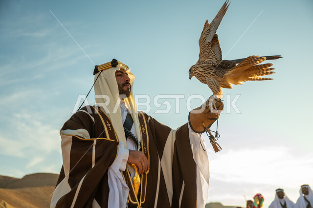 يوم بدينا 22 فبراير، الاهتمام بتربية الطيور و تدريبها, مكانة الصقور في التاريخ السعودي, صورة مقربة لرجل عربي خليجي سعودي يرتدي الغترة والبشت يحمل على يده طائر الصقر, ذكرى تاسيس الدوله السعوديه الاولى 1727 م, مناسبة وطنيه شعبيه, خلفية العرضة النجدية  في الصحراء