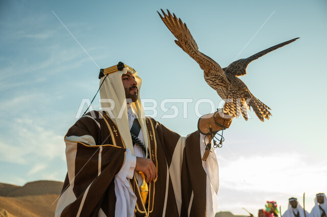 يوم بدينا 22 فبراير، الاهتمام بتربية الطيور و تدريبها, مكانة الصقور في التاريخ السعودي, صورة مقربة لرجل عربي خليجي سعودي يرتدي الغترة والبشت يحمل على يده طائر الصقر, ذكرى تاسيس الدوله السعوديه الاولى 1727 م, مناسبة وطنيه شعبيه, خلفية العرضة النجدية  في الصحراء