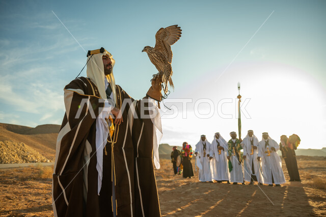 ذكرى تاسيس الدوله السعوديه الاولى 1727 م, مكانة الصقور في التاريخ السعودي, مناسبة شعبيه وطنيه, يوم بدينا 22 فبراير، الاهتمام بتربية الطيور و تدريبها, رجل عربي خليجي سعودي يرتدي الغترة والبشت يحمل على يده طائر الصقر بتعابير الفخر, خلفية العرضة النجدية في الصحراء