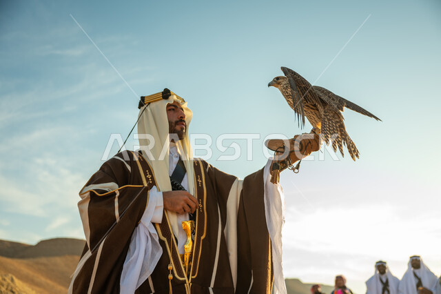 مناسبة وطنيه شعبيه, ذكرى تاسيس الدوله السعوديه الاولى 1727 م, يوم بدينا 22 فبراير، الاهتمام بتربية الطيور و تدريبها, مكانة الصقور في التاريخ السعودي, صورة مقربة لرجل عربي خليجي سعودي يرتدي الغترة والبشت يحمل على يده طائر الصقر, خلفية العرضة النجدية في الصحراء