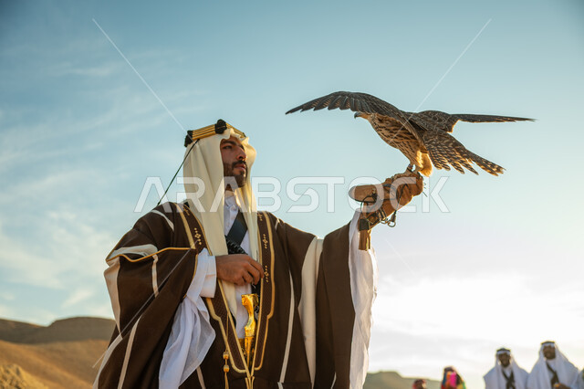 مناسبة وطنيه شعبيه, ذكرى تاسيس الدوله السعوديه الاولى 1727 م, يوم بدينا 22 فبراير، الاهتمام بتربية الطيور و تدريبها, مكانة الصقور في التاريخ السعودي, صورة مقربة لرجل عربي خليجي سعودي يرتدي الغترة والبشت يحمل على يده طائر الصقر, خلفية العرضة النجدية في الصحراء