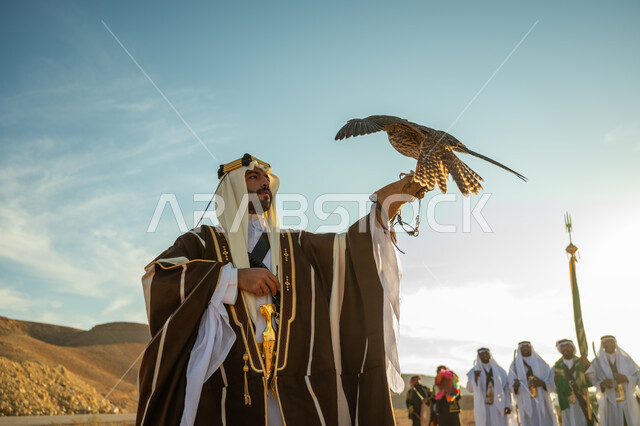 العناية بتربية الطيور و تدريبها, مكانة الصقور في التاريخ السعودي, ذكرى تاسيس الدوله السعوديه الاولى 1727 م, يوم بدينا 22 فبراير، مناسبة وطنيه شعبيه, رجل عربي خليجي سعودي يرتدي الغترة والبشت يحمل على يده طائر الصقر بتعابير الفخر, خلفية العرضة النجدية في الصحراء