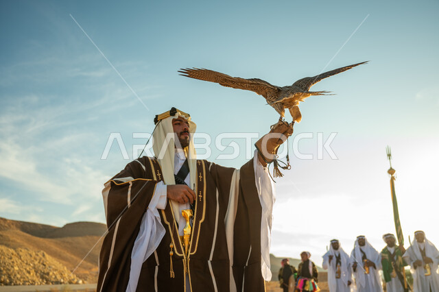 مكانة الصقور في التاريخ السعودي, ذكرى تاسيس الدوله السعوديه الاولى 1727 م, يوم بدينا 22 فبراير، مناسبة وطنيه شعبيه, الاهتمام بتربية الطيور و تدريبها, رجل عربي خليجي سعودي يرتدي الغترة والبشت يحمل على يده طائر الصقر بتعابير الفخر, خلفية العرضة النجدية في الصحراء