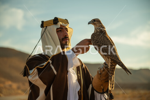 مناسبة وطنيه شعبيه ,الاهتمام بتربية الطيور و تدريبها, مكانة الصقور في التاريخ السعودي, صورة مقربة لرجل عربي خليجي سعودي يرتدي الغترة والبشت يحمل على يده طائر الصقر بتعابير الفخر في الصحراء, يوم بدينا 22 فبراير، ذكرى تاسيس الدوله السعوديه الاولى 1727 م