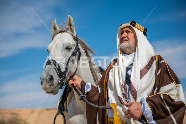 The day we started is February 22, the inheritance of the love and breeding of horses from our ancestors, a close-up of a Saudi Arabian Gulf man wearing a golden headband and a bisht standing next to his purebred white horse, the anniversary of the founding of the first Saudi state in 1727 AD, wearing traditional dress on a national occasion, caring for horses in the desert