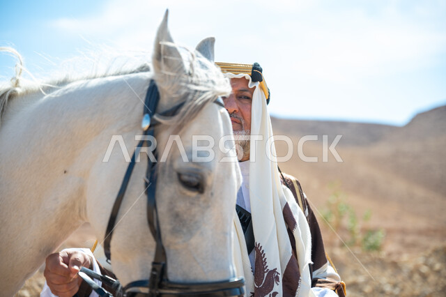 توراث حب وتربية الجياد من الأجداد, صورة مقربة لرجل عربي خليجي سعودي يرتدي العقال الذهبي والبشت يقف بجانب فرسه الأبيض الأصيل, يوم بدينا  22 فبراير, ذكرى تأسيس الدولة السعودية الأولى في 1727م, ارتداء الزي الشعبي في المناسبة الوطنية، رعاية الخيل في الصحراء