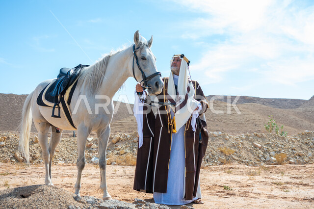 ذكرى تأسيس الدولة السعودية الأولى في 1727م, توراث حب وتربية الجياد من الأجداد, رعاية الخيل والاهتمام به في الصحراء، رجل عربي خليجي سعودي يرتدي العقال الذهبي والبشت يقف بجانب فرسه الأبيض الأصيل, ارتداء الزي الشعبي في المناسبة الوطنية، يوم بدينا  22 فبراير