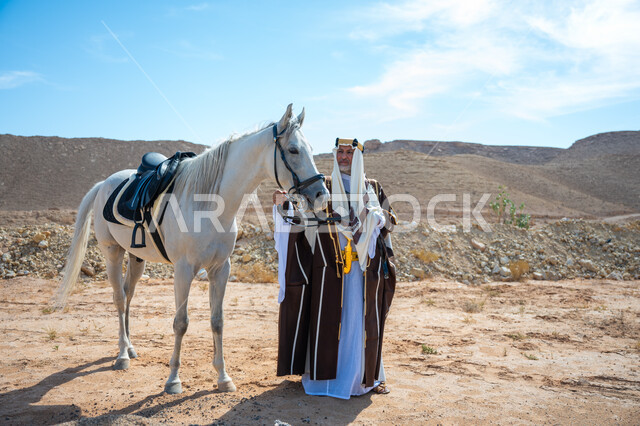 Inheriting the love and breeding of horses from our ancestors, caring for and looking after horses in the desert, a Saudi Arabian Gulf man wearing a golden headband and a bisht standing next to his purebred white horse, wearing traditional dress on a national occasion, the anniversary of the founding of the first Saudi state in 1727 AD, the day we started, February 22