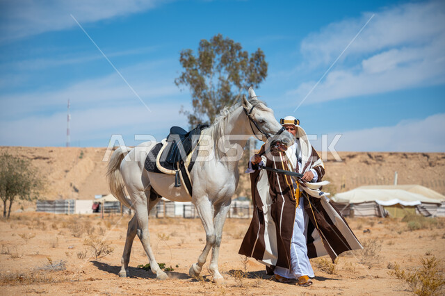 يوم بدينا  22 فبراير، رعاية الخيل والاهتمام به في الصحراء، رجل عربي خليجي سعودي يرتدي العقال الذهبي والبشت يمشي برفقة فرسه الأبيض الأصيل, ارتداء الزي الشعبي في المناسبة الوطنية، ذكرى تأسيس الدولة السعودية الأولى في 1727م, توراث حب وتربية الجياد من الأجداد
