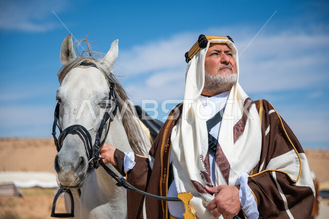 The day we started is February 22, the inheritance of the love and breeding of horses from our ancestors, a close-up of a Saudi Arabian Gulf man wearing a golden headband and a bisht standing next to his purebred white horse, the anniversary of the founding of the first Saudi state in 1727 AD, wearing traditional dress on a national occasion, caring for horses in the desert