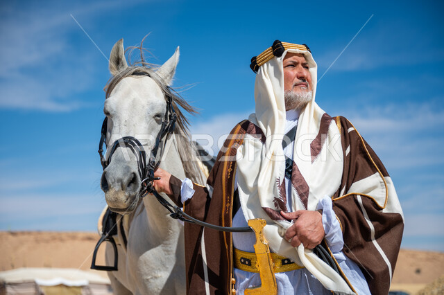 ذكرى تأسيس الدولة السعودية الأولى في 1727م, يوم بدينا  22 فبراير, توراث حب وتربية الجياد من الأجداد, صورة مقربة لرجل عربي خليجي سعودي يرتدي العقال الذهبي والبشت يقف بجانب فرسه الأبيض الأصيل, ارتداء الزي الشعبي في المناسبة الوطنية، رعاية الخيل في الصحراء