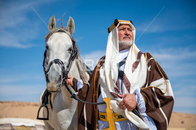 ذكرى تأسيس الدولة السعودية الأولى في 1727م, يوم بدينا  22 فبراير, توراث حب وتربية الجياد من الأجداد, صورة مقربة لرجل عربي خليجي سعودي يرتدي العقال الذهبي والبشت يقف بجانب فرسه الأبيض الأصيل, ارتداء الزي الشعبي في المناسبة الوطنية، رعاية الخيل في الصحراء