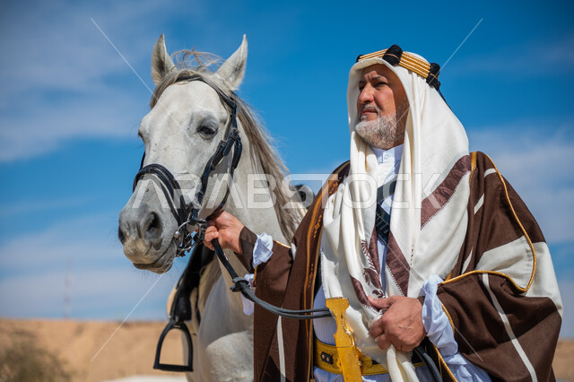 ارتداء الزي الشعبي في المناسبة الوطنية، ذكرى تأسيس الدولة السعودية الأولى في 1727م, يوم بدينا  22 فبراير, توراث حب وتربية الجياد من الأجداد, صورة مقربة لرجل عربي خليجي سعودي يرتدي العقال الذهبي والبشت يقف بجانب فرسه الأبيض الأصيل, رعاية الخيل في الصحراء