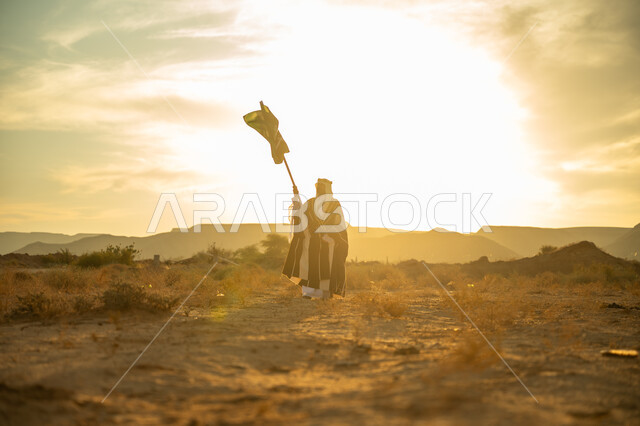 The day we started is February 22, the anniversary of the founding of the first Saudi state in 1727 AD, a special national holiday and occasion, wearing the central region costume, pride and honor in the national identity, a Saudi Gulf Arab man wearing the bisht and the golden agal raising the flagpole of the Kingdom at sunrise in the desert