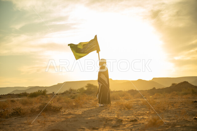 الاعتزاز والفخر بالهوية الوطنية, ارتداء زي المنطقة الوسطى, مناسبة وعيد وطني متميز, الاحتفال بذكرى تأسيس الدولة السعودية الأولى 1727م, يوم بدينا 22 فبراير, رجل عربي خليجي سعودي يرتدي البشت والعقال يرفع سارية علم المملكة عند شروق الشمس في الصحراء