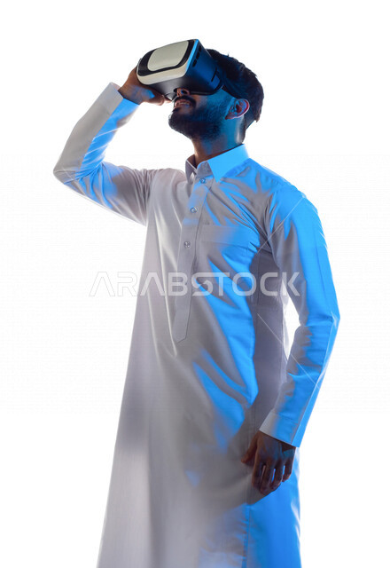 بورتريه لرجل عربي خليجي سعودي مبتسم يرتدي نظارة الواقع الإفتراضي ثلاثية الأبعاد الفريدة و العجيبة و ينظر إلى أعلى ، التفاعل و المتعة و التسلية بمشاهدة الأفلام و محاكاة العالم الإفتراضي ، خلفية بيضاء