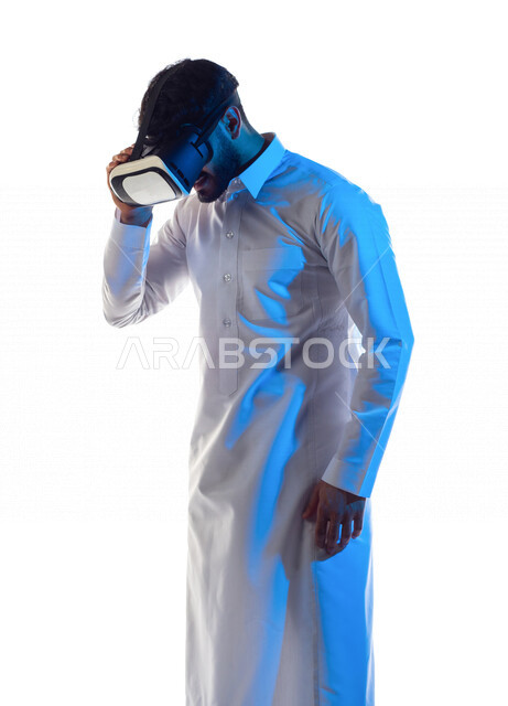 بورتريه لرجل عربي خليجي سعودي مبتسم يرتدي نظارة الواقع الإفتراضي ثلاثية الأبعاد الفريدة و العجيبة و ينظر إلى الأسفل ، التفاعل و المتعة و التسلية بمشاهدة الأفلام و محاكاة العالم الإفتراضي ، خلفية بيضاء