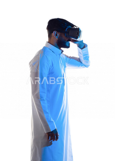 بورتريه من الجانب لرجل عربي خليجي سعودي مبتسم  يرتدي نظارة الواقع الإفتراضي ثلاثية الأبعاد الفريدة و العجيبة و يمسكها بيده ، التفاعل و المتعة و التسلية بمشاهدة الأفلام و محاكاة العالم الإفتراضي ، خلفية بيضاء