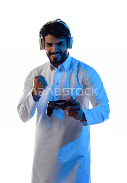 بورتريه لرجل عربي خليجي سعودي مبتسم ، يرتدي سماعة رأس و يمسك بيده عصا التحكم و يلعب بألعاب الفيديو ، بإيماءات و تعابير تدل على الفوز ، المتعة و التسلية بألعاب الفيديو ، ألعاب البلايستيشن الترفيهية ، تقنيات و ألعاب