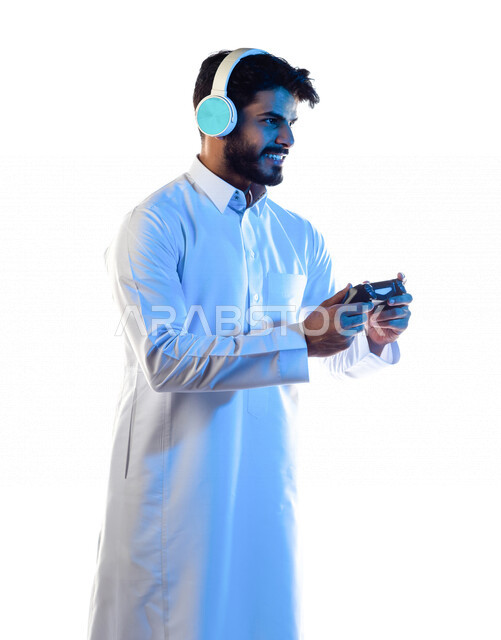 بورتريه لرجل عربي خليجي سعودي مبتسم ، يرتدي سماعة رأس و يمسك عصا التحكم و يلعب بألعاب الفيديو ، المتعة و التسلية بألعاب الفيديو و ألعاب العالم الافتراضي ، ألعاب البلايستيشن الترفيهية ، تقنيات و ألعاب ، خلفية بيضاء