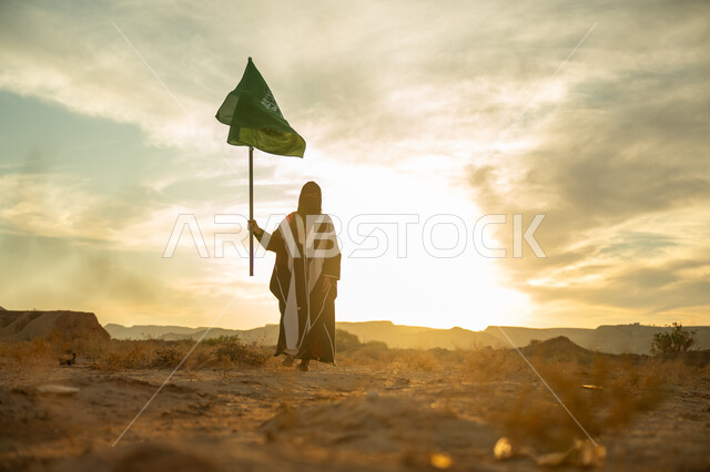 يوم بدينا 22 فبراير, الاحتفال بذكرى تأسيس الدولة السعودية الأولى 1727م, ارتداء الزي الشعبي التقليدي في مناسبة وطنية, حب الوطن والانتماء اليه, امرأة عربية خليجي سعودية ترتدي البرقع والعباءة ترفع سارية علم المملكة عند شروق الشمس في الطبيعة الصحراوية