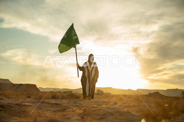 ذكرى تأسيس الدولة السعودية الأولى 1727م, ارتداء الزي الشعبي التقليدي في مناسبة وطنية, حب الوطن والانتماء اليه, يوم بدينا 22 فبراير, امرأة عربية خليجي سعودية ترتدي البرقع والعباءة ترفع سارية علم المملكة عند شروق الشمس في الطبيعة الصحراوية