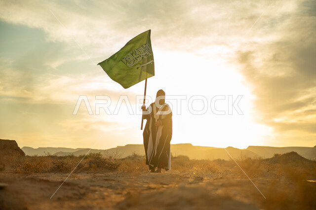 يوم بدينا 22 فبراير, الاحتفال بذكرى تأسيس الدولة السعودية الأولى 1727م, ارتداء الزي الشعبي التقليدي في مناسبة وطنية, الاعتزاز والفخر بالانتماء للوطن, امرأة عربية خليجي سعودية ترتدي البرقع والعباءة ترفع سارية علم المملكة عند شروق الشمس في الطبيعة الصحراوية