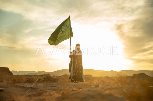 الاحتفال بذكرى تأسيس الدولة السعودية الأولى 1727م, ارتداء الزي الشعبي التقليدي في مناسبة وطنية, الاعتزاز والفخر بالانتماء للوطن, يوم بدينا 22 فبراير, امرأة عربية خليجي سعودية ترتدي البرقع والعباءة ترفع سارية علم المملكة عند شروق الشمس في الطبيعة الصحراوية