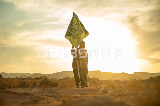 ذكرى تأسيس الدولة السعودية الأولى 1727م, ارتداء الزي الشعبي التقليدي في مناسبة وطنية, الاعتزاز والفخر بالانتماء للوطن, يوم بدينا 22 فبراير, امرأة عربية خليجي سعودية ترتدي البرقع والعباءة ترفع سارية علم المملكة عند شروق الشمس في الطبيعة الصحراوية