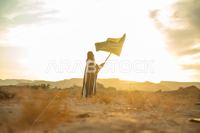 الفخر والاعتزاز بالهوية الوطنية, يوم بدينا 22 فبراير, ذكرى تأسيس الدولة السعودية الأولى 1727م, ارتداء الزي الشعبي التقليدي في مناسبة وطنية, صورة من الجانب لامرأة عربية خليجي سعودية ترتدي البرقع والعباءة ترفع سارية علم المملكة عند شروق الشمس في الطبيعة الصحراوية