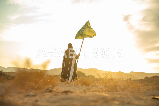 يوم بدينا 22 فبراير, ذكرى تأسيس الدولة السعودية الأولى 1727م, ارتداء الزي الشعبي التقليدي في مناسبة وطنية, الاعتزاز والفخر بالهوية الوطنية, صورة من الخلف لامرأة عربية خليجي سعودية ترتدي البرقع والعباءة ترفع سارية علم المملكة عند شروق الشمس في الطبيعة الصحراوية