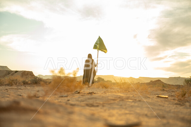 ذكرى تأسيس الدولة السعودية الأولى 1727م, ارتداء الزي الشعبي التقليدي في مناسبة وطنية, الاعتزاز والفخر بالهوية الوطنية, يوم بدينا 22 فبراير, صورة من الخلف لامرأة عربية خليجي سعودية ترتدي البرقع والعباءة ترفع سارية علم المملكة عند شروق الشمس في الطبيعة الصحراوية