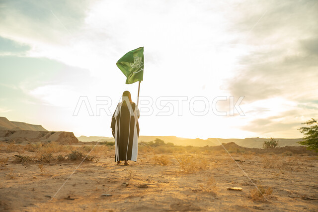 The day we started is February 22, the anniversary of the founding of the first Saudi state in 1727 AD, wearing traditional folk costume on a national occasion, the concept of love of the homeland and belonging to it, a picture from the back of a Saudi Arabian Gulf woman wearing a burqa and an abaya raising the flagpole of the Kingdom at sunrise in the desert nature