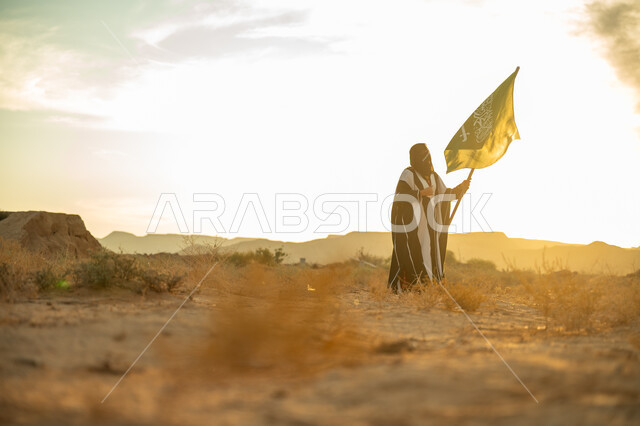 ذكرى تأسيس الدولة السعودية الأولى 1727م, ارتداء الزي الشعبي التقليدي في مناسبة وطنية, الاعتزاز والفخر بالانتماء للوطن, يوم بدينا 22 فبراير, امرأة عربية خليجي سعودية ترتدي البرقع والعباءة ترفع سارية علم المملكة عند شروق الشمس في الطبيعة الصحراوية