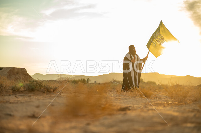 الاحتفال بذكرى تأسيس الدولة السعودية الأولى 1727م, ارتداء الزي الشعبي التقليدي في مناسبة وطنية, الاعتزاز والفخر بالانتماء للوطن, يوم بدينا 22 فبراير, امرأة عربية خليجي سعودية ترتدي البرقع والعباءة ترفع سارية علم المملكة عند شروق الشمس في الطبيعة الصحراوية