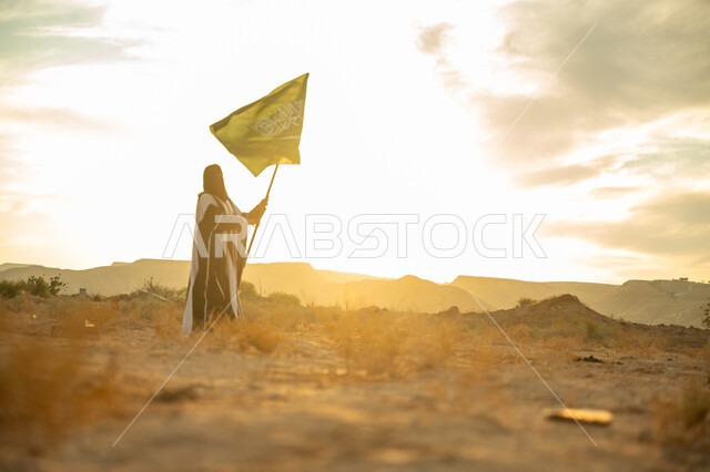 الفخر والاعتزاز بالانتماء للوطن, يوم بدينا 22 فبراير, الاحتفال بذكرى تأسيس الدولة السعودية الأولى 1727م, ارتداء الزي الشعبي التقليدي في مناسبة وطنية, امرأة عربية خليجي سعودية ترتدي البرقع والعباءة ترفع سارية علم المملكة عند شروق الشمس في الطبيعة الصحراوية