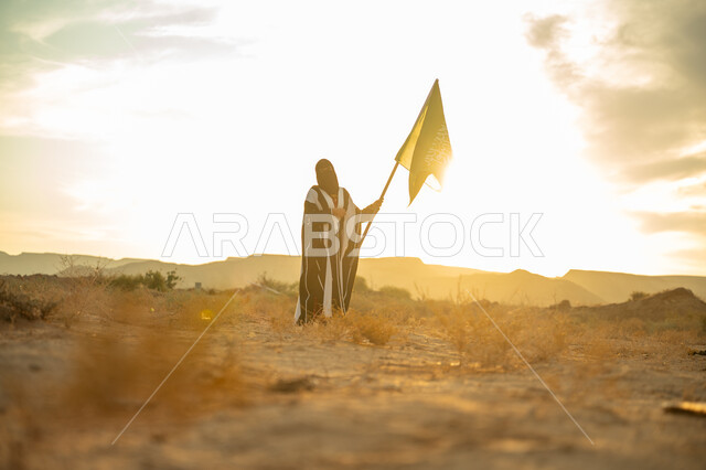 Celebrating the anniversary of the founding of the first Saudi state 1727 AD, wearing traditional folk costume on a national occasion, the concept of love of the homeland and belonging to it, the day we started on February 22, a Saudi Arabian Gulf woman wearing a burqa and an abaya raises the flagpole of the Kingdom at sunrise in the desert nature