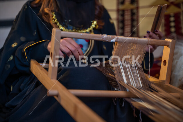 ذكرى تأسيس الدولة السعودية الأولى في 1727م، صورة مقربة لامرأة عربية خليجية سعودية ترتدي العباءة والبرقع تقوم بحياكة وغزل الأنسجة والقماش, مهنة تقليدية قديمة, يوم بدينا  22 فبراير، حرفة يدوية تاريخية, منتج محلي وطني، ارتداء الزي الشعبي في المناسبة الوطنية