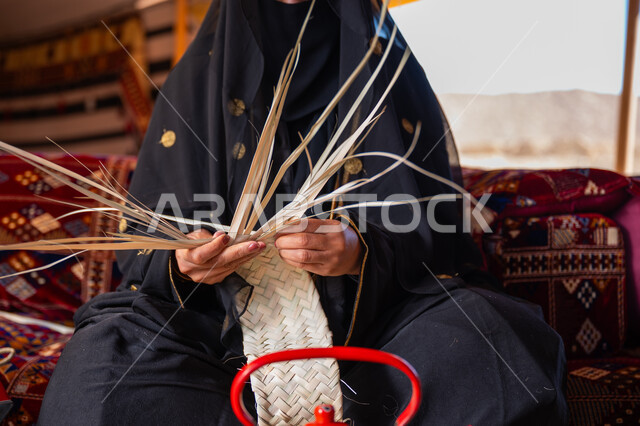 Using dried palm leaves in basket making, a traditional handicraft, the anniversary of the founding of the first Saudi state in 1727 AD, a close-up of a Saudi Gulf Arab woman wearing a niqab and an abaya sitting in a tent in the desert weaving palm fronds, wearing traditional dress on the national occasion, the day of our beginning, February 22