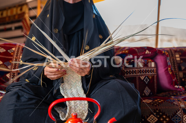 Wearing traditional costume on a national occasion, using dried palm leaves in making baskets, a traditional handicraft, the anniversary of the founding of the first Saudi state in 1727 AD, a close-up of a Saudi Gulf Arab woman wearing a niqab and an abaya sitting in a tent in the desert braiding palm fronds, the day we started on February 22