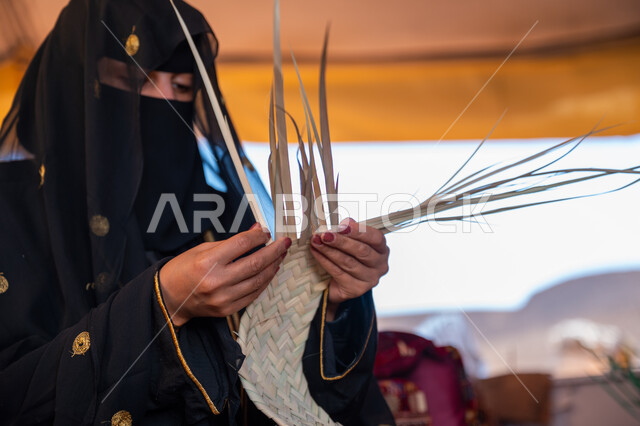 ذكرى تأسيس الدولة السعودية الأولى في 1727م، صورة مقربة لامرأة عربية خليجية سعودية ترتدي النقاب والعباءة تجلس في خيمة في الصحراء تقوم بجدل سعائف الخوص, حرفة يدوية تقليدية, يوم بدينا 22 فبراير، ارتداء الزي الشعبي في المناسبة الوطنية، استخدام أوراق النخيل المجففة في صناعة السلل