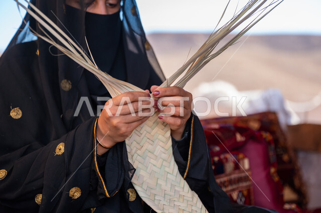 استخدام أوراق النخيل المجففة في صناعة السلل، حرفة يدوية تقليدية, ذكرى تأسيس الدولة السعودية الأولى في 1727م، صورة مقربة لامرأة عربية خليجية سعودية ترتدي النقاب والعباءة تجلس في خيمة في الصحراء تقوم بجدل سعائف الخوص, ارتداء الزي الشعبي في المناسبة الوطنية، يوم بدينا 22 فبراير