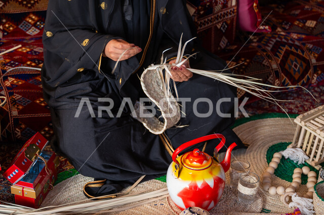 ارتداء الزي الشعبي في المناسبة الوطنية، استخدام أوراق النخيل المجففة في صناعة السلل، حرفة يدوية تقليدية, ذكرى تأسيس الدولة السعودية الأولى في 1727م، صورة مقربة لامرأة عربية خليجية سعودية ترتدي العباءة تجلس في خيمة في الصحراء تقوم بجدل سعائف الخوص, يوم بدينا 22 فبراير