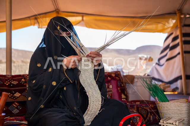 Traditional handicraft, commemoration of the founding of the first Saudi state in 1727 AD, a Saudi Arabian Gulf woman wearing a niqab and abaya sitting in a tent in the desert weaving palm fronds, wearing traditional dress on the national occasion, the day of our beginning, February 22, using dried palm leaves in making baskets
