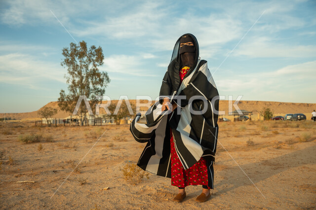 ذكرى تأسيس الدولة السعودية الأولى 1727 م، ارتداء زي المنطقة الوسطى في المناسبة الوطنية، الاهتمام والعناية بالمظهر الخارجي، إمرأة خليجية عربية سعودية ترتدي البشت النسائي السعودي والبرقع تقف على الرمال الذهبية الناعمة في الصحراء، يوم بدينا 22 فبراير