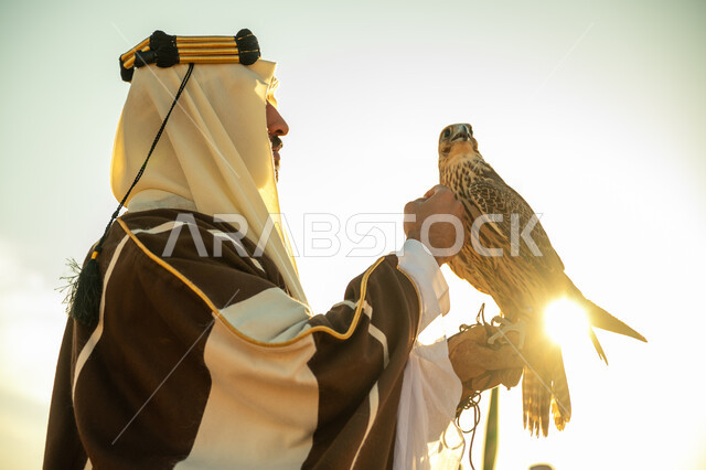 Interest in raising and training birds, the status of falcons in Saudi history, the anniversary of the founding of the first Saudi state 1727 AD, the day we started February 22, a close-up photo from the side of a Saudi Gulf Arab man wearing a ghutra and a bisht holding a falcon in his hand with expressions of pride, a background of the sky with the bright sun